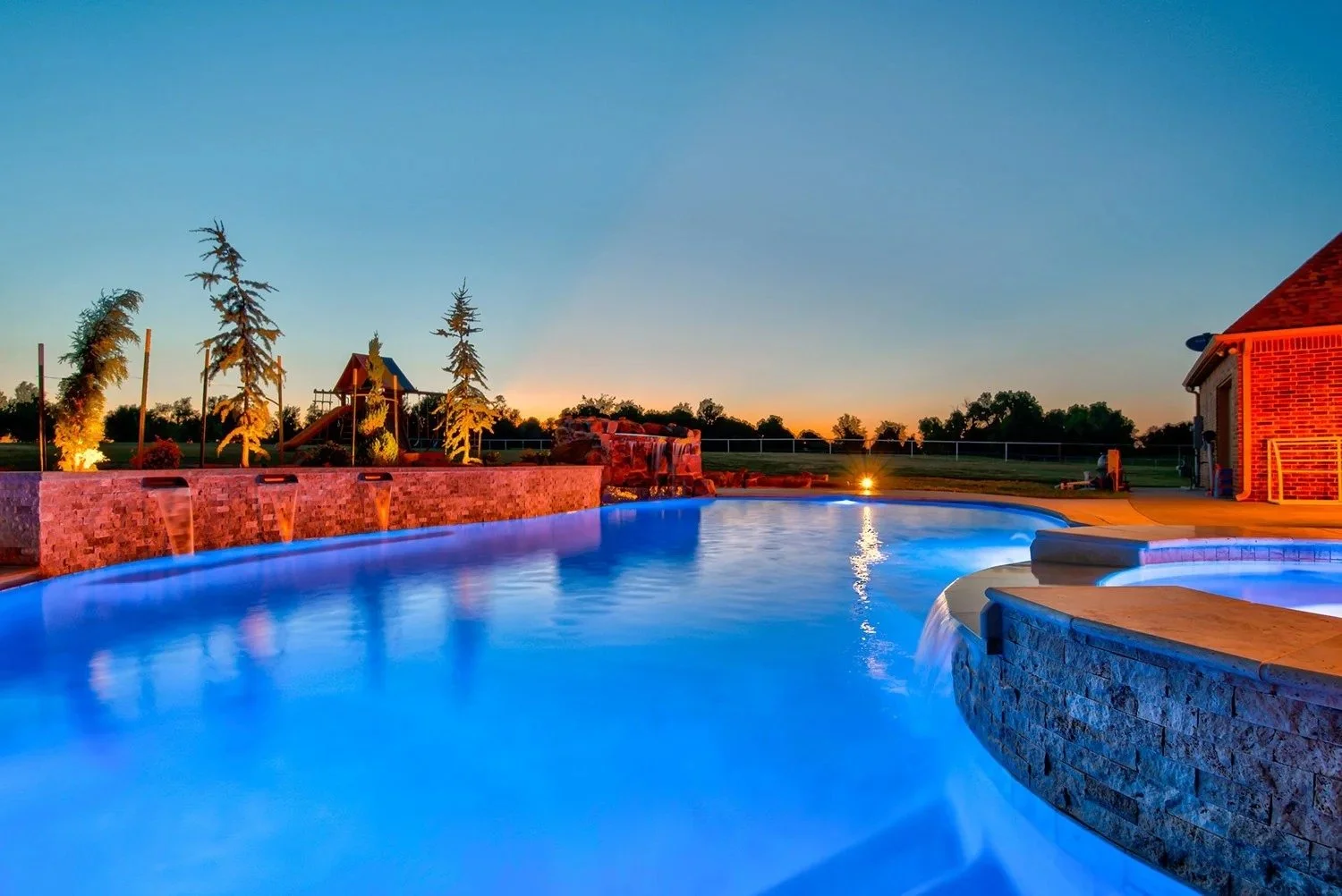 An illuminated backyard swimming pool at dusk with water features, surrounded by a brick patio, tall trees, a play structure, a brick house, and a sunset sky.