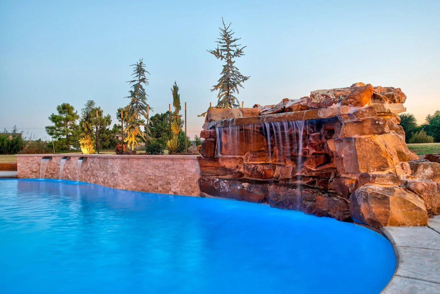 A swimming pool with water features and a red rock waterfall structure surrounded by trees at sunset.