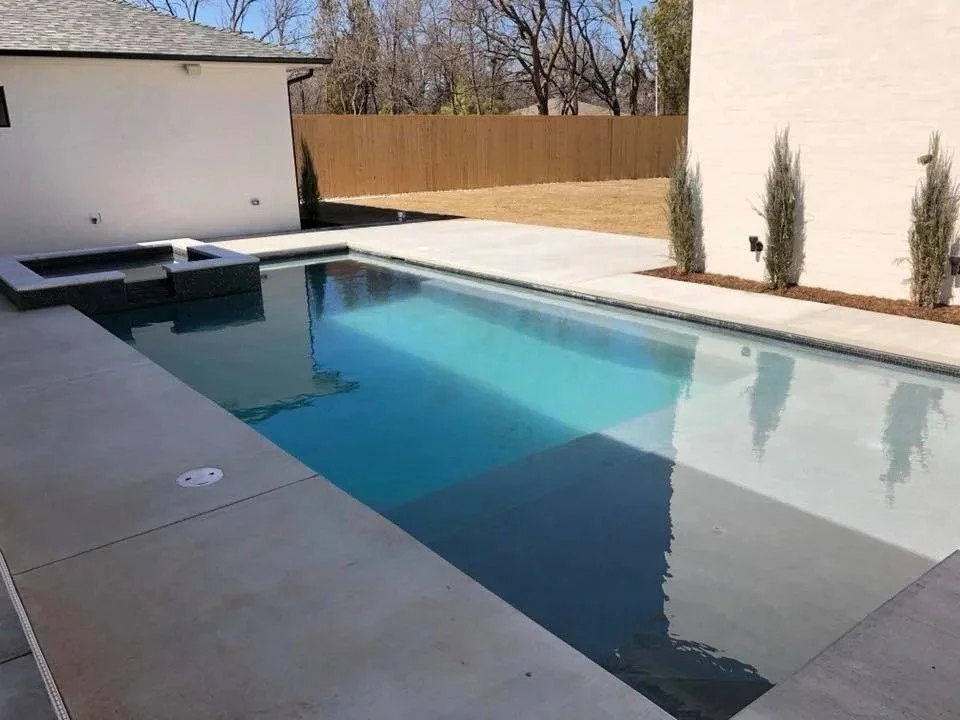 A modern backyard with a rectangular in-ground swimming pool, a small hot tub, and a concrete patio. There are three small trees along the fence, and the yard is surrounded by a tall wooden privacy fence.