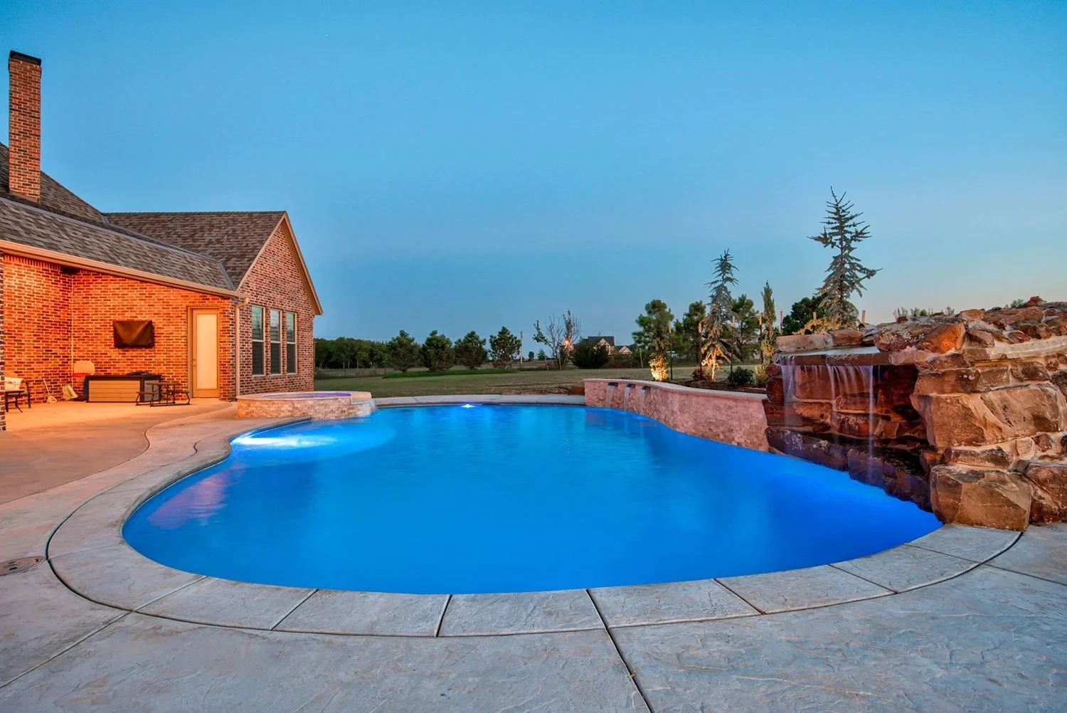 A backyard swimming pool with blue water, surrounded by a concrete deck, a brick house, and a stone waterfall feature during dusk.