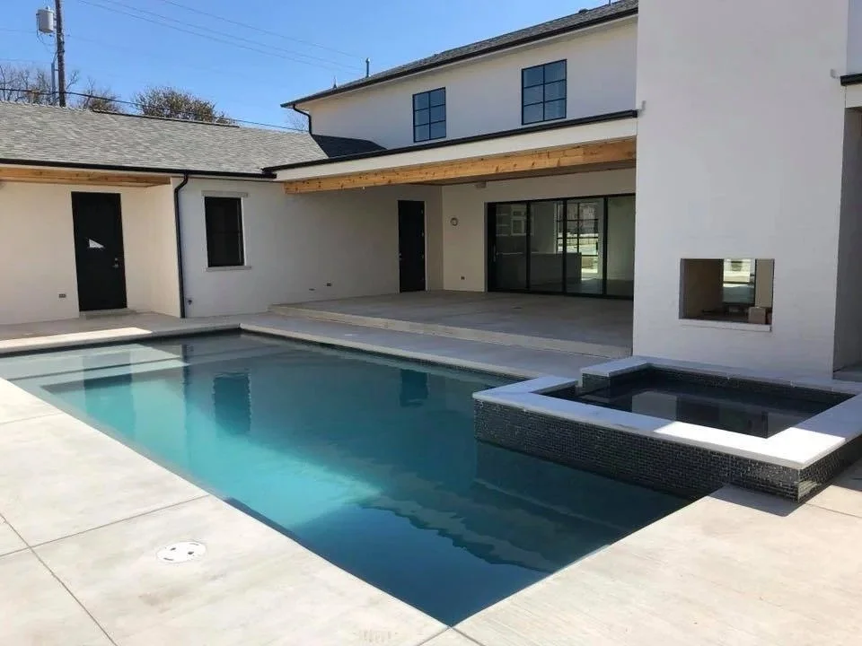 Modern backyard with an in-ground swimming pool, hot tub, and patio area, surrounded by a white house with large windows.