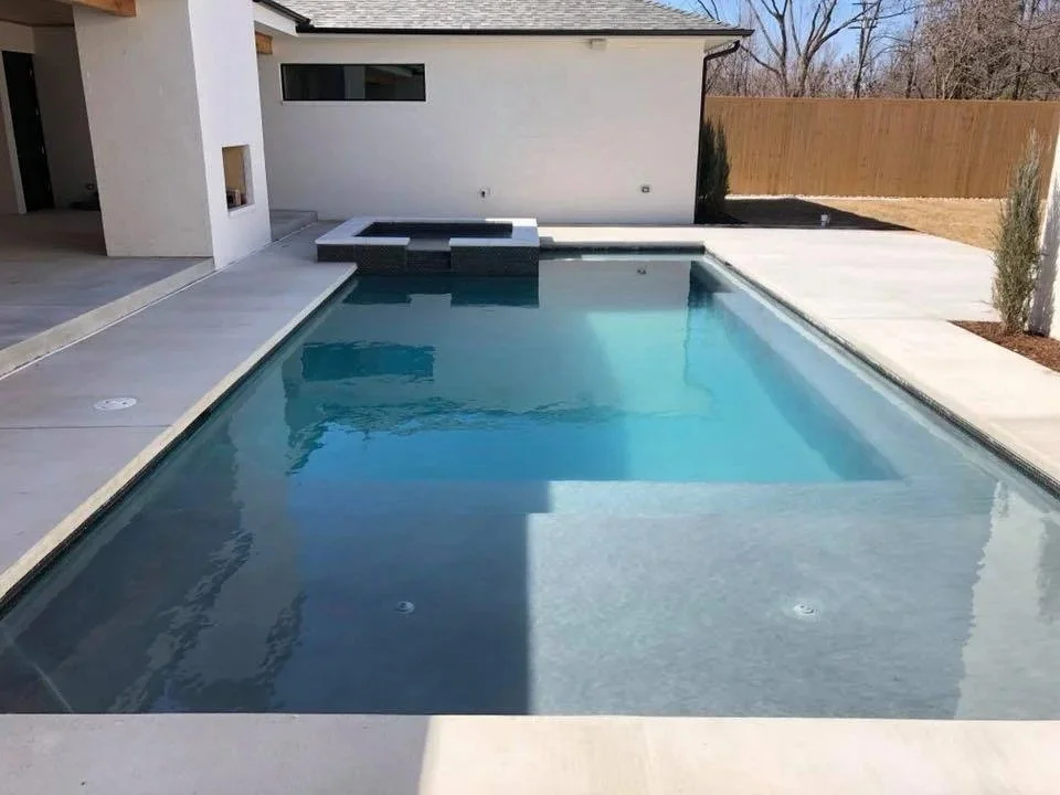 A backyard with a rectangular in-ground swimming pool, a white building with a small window, and a wooden fence. There are two small trees and a paved patio area.