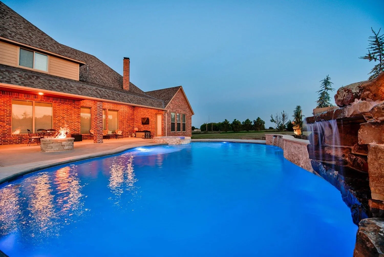 A backyard with a swimming pool, a fire pit, and a stone waterfall feature, with a brick house in the background during twilight.