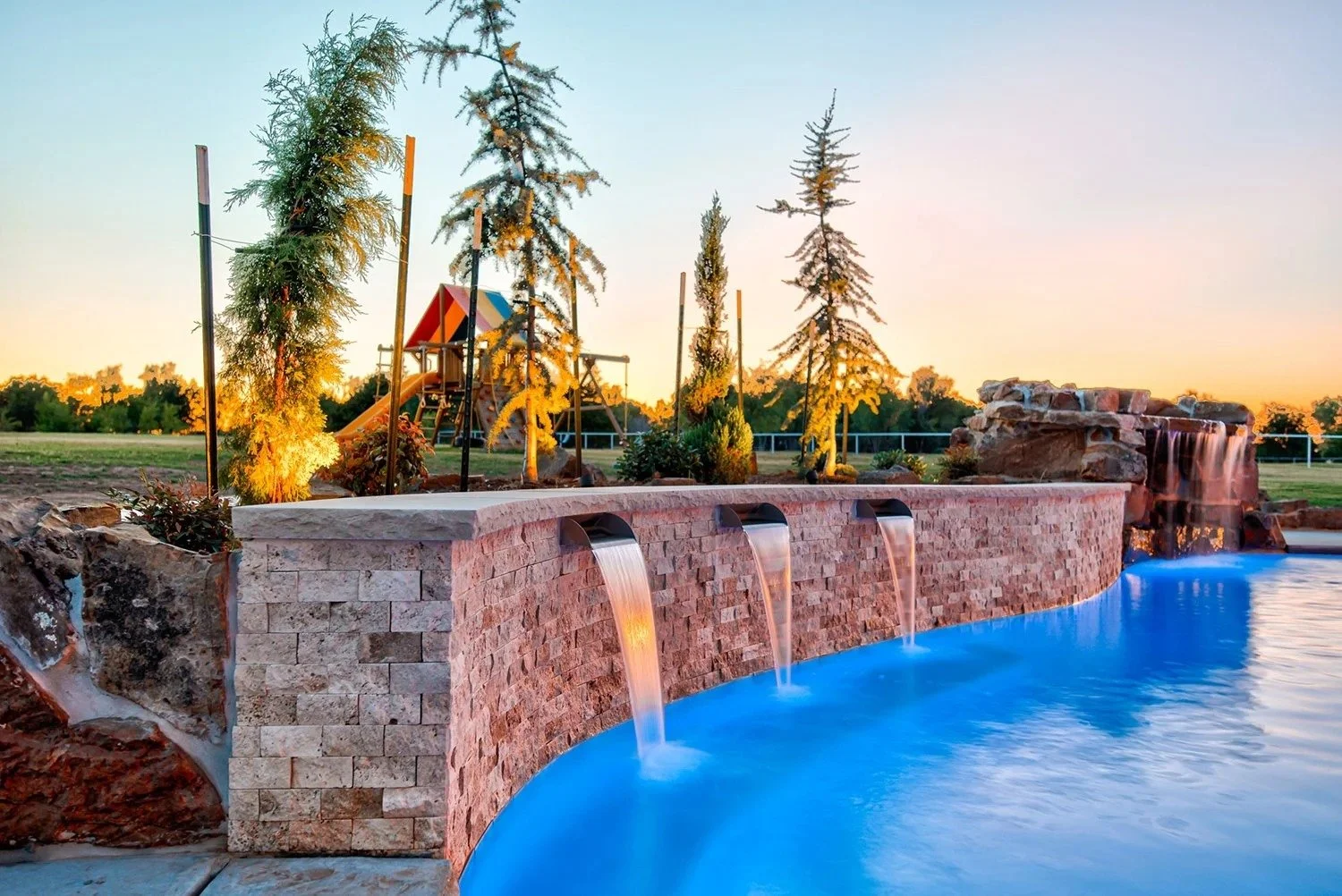 A backyard pool area at sunset with a stone waterfall feature, a brick wall with water spouts, trees, and a playground in the background.
