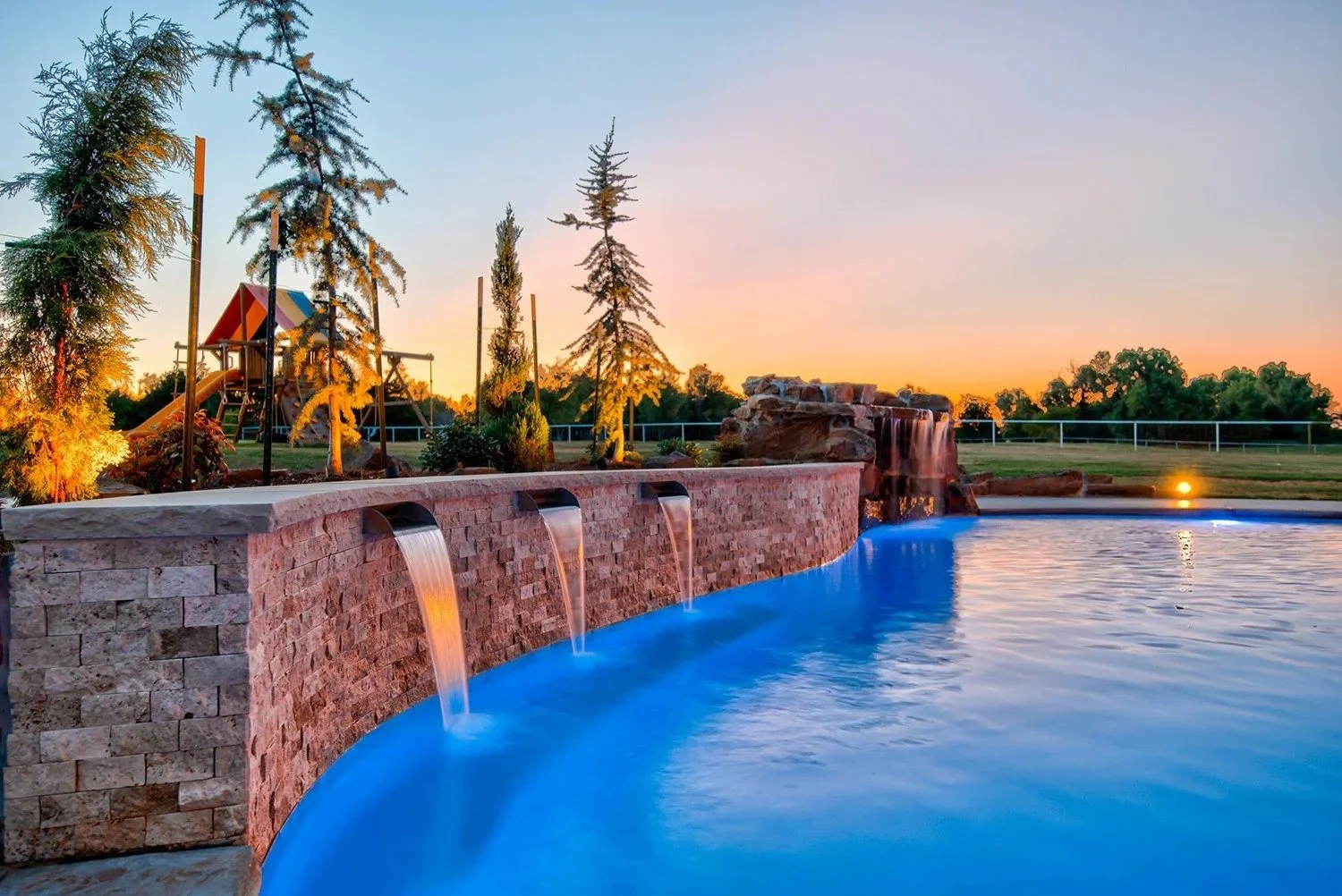 An outdoor swimming pool at sunset with a brick waterfall feature and a water slide in the background, surrounded by trees and a grassy field.
