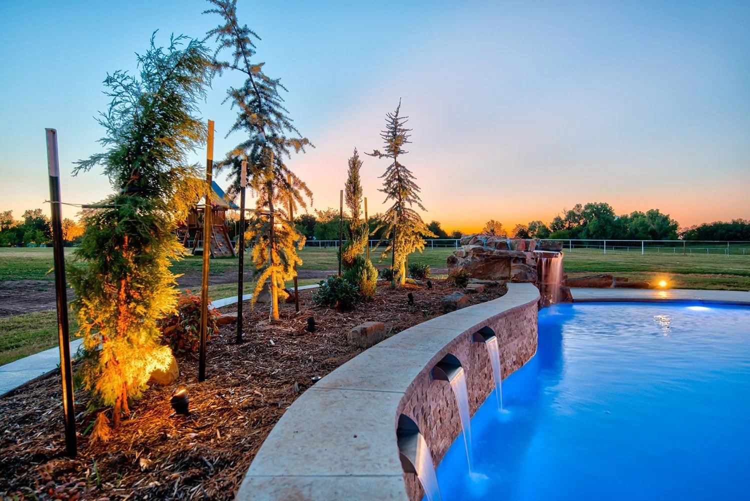 Outdoor pool with a water feature, surrounded by landscaped plants and trees, at sunset.