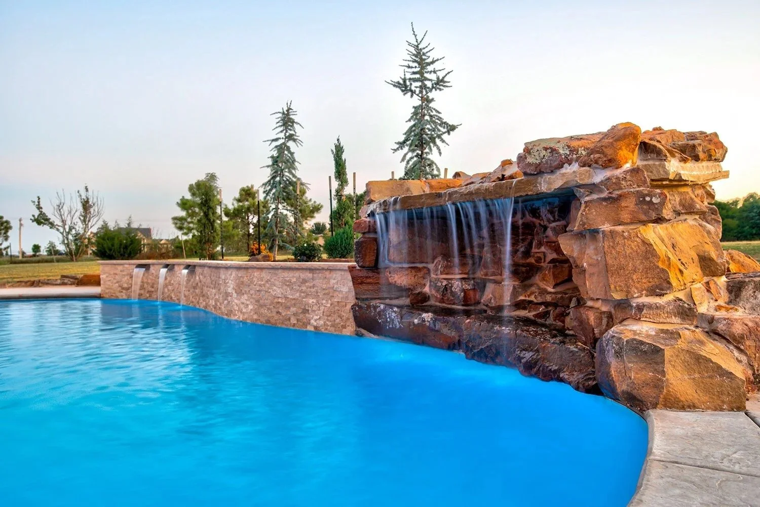 A backyard swimming pool with a stone waterfall feature and lush trees in the background.