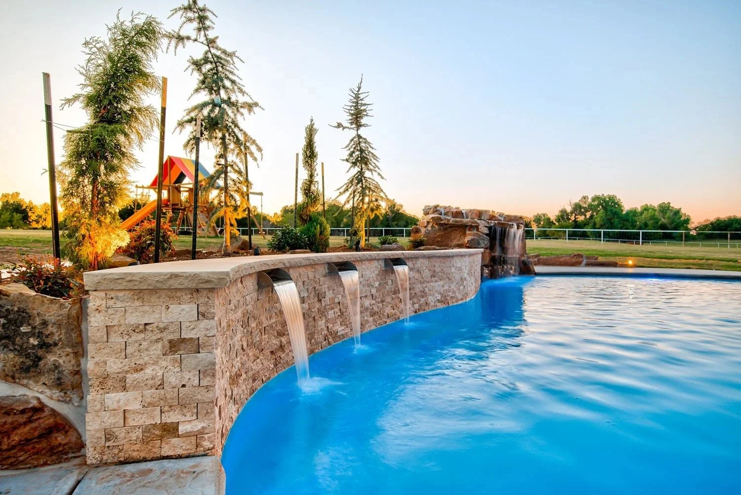 An outdoor pool with water features, a waterfall, and a stone deck. In the background, there are trees, a grassy field, and a playground with a slide, under a clear sky during sunset.