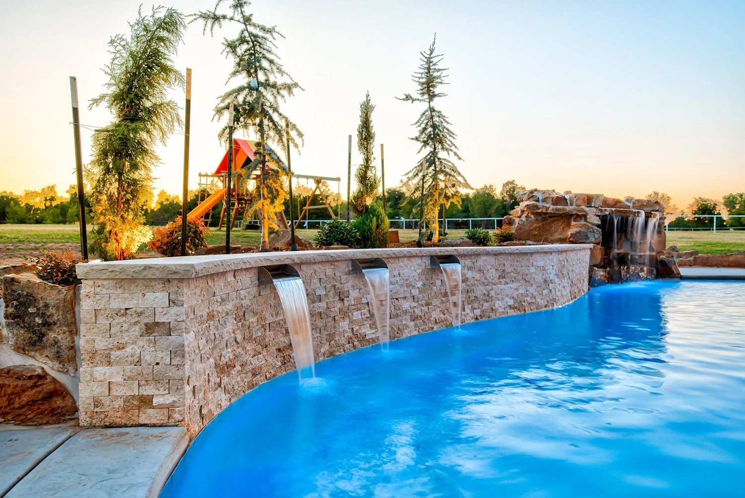 A backyard with a stone waterfall feature flowing into a pool, with trees and a children's playground with a slide in the background at sunset.
