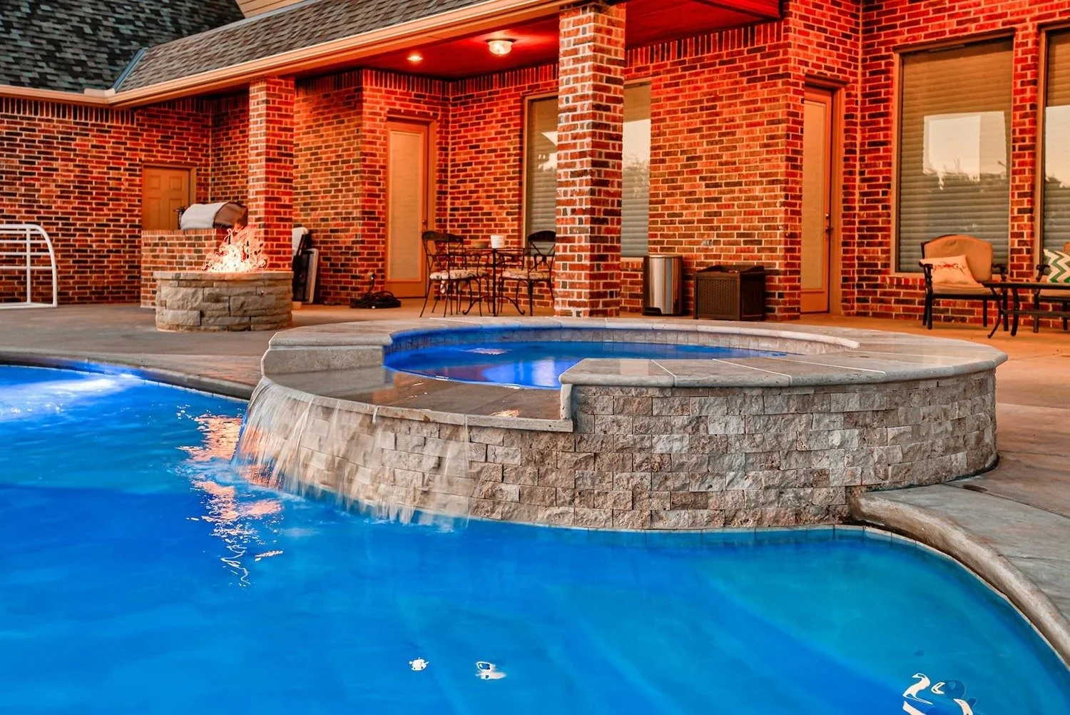 Backyard with a swimming pool, hot tub, brick house, outdoor furniture, fire pit, and lounge chair.