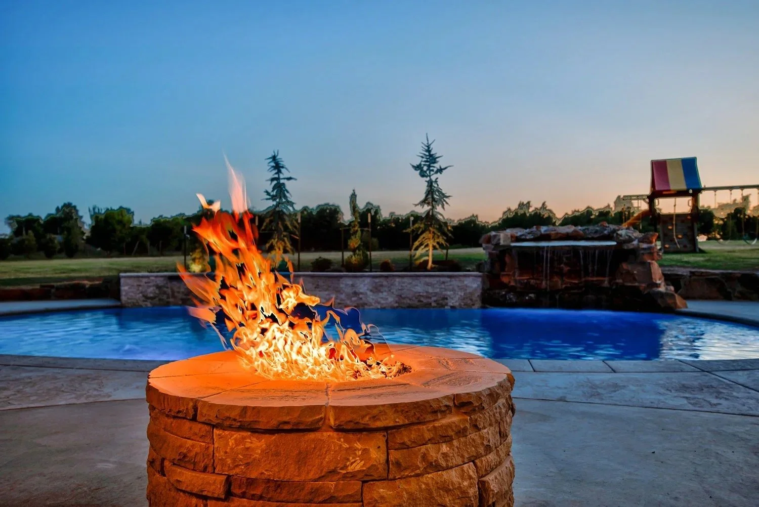 A backyard swimming pool with a stone fire pit in the foreground, a waterfall feature, and a playground with swings and a slide in the background at sunset.