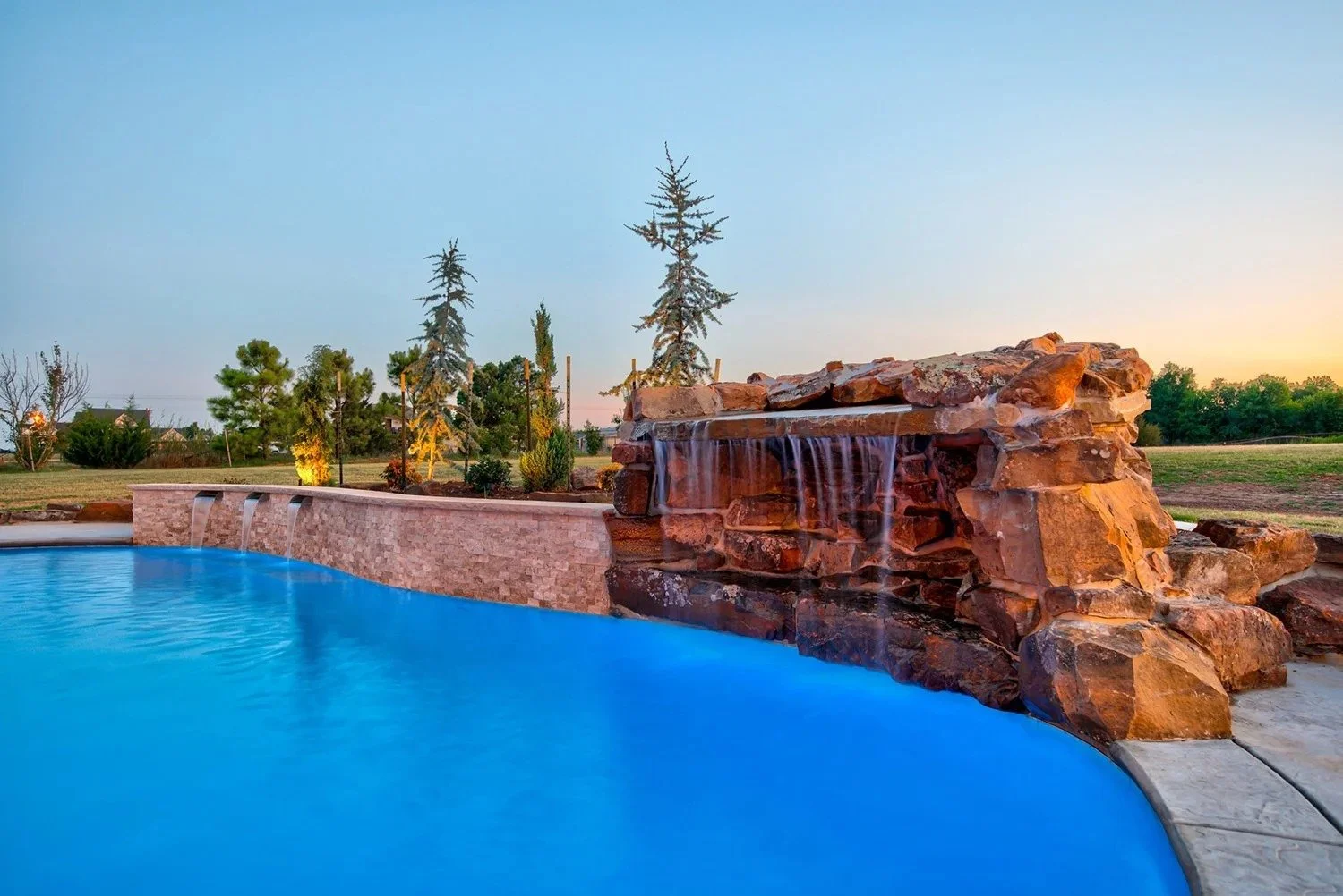 An outdoor swimming pool with a rock waterfall feature, surrounded by a brick wall and trees at sunset.