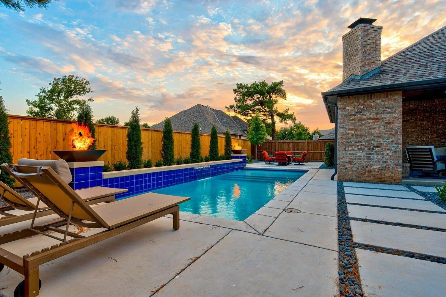 Backyard with a swimming pool, lounge chairs, fire bowls, and outdoor seating, surrounded by a wooden fence and trees at sunset.