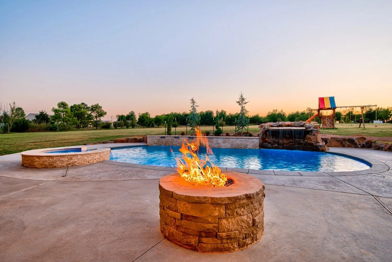 A backyard with a curved swimming pool, a fire pit with a flame, a water feature with a waterfall, and a playground with a slide and swings at sunset.