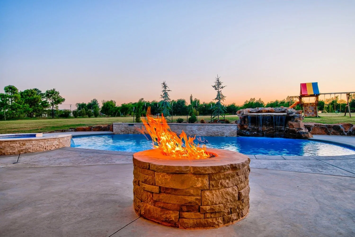 Backyard with a swimming pool, fire pit, waterfall feature, and playground area at sunset.