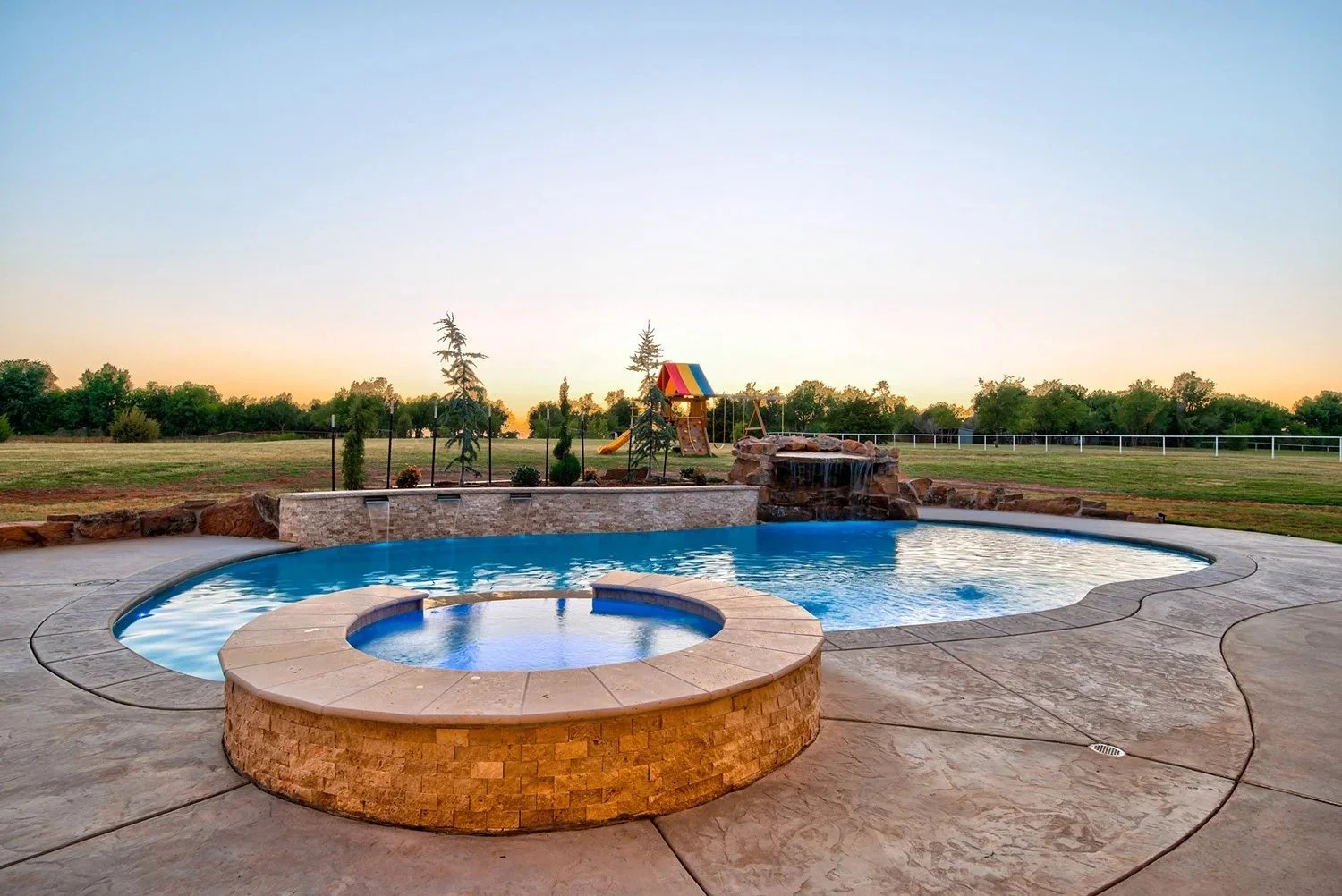 A backyard swimming pool with a hot tub, waterfall feature, and a playground with a slide and swing set, surrounded by a large grassy yard and trees at sunset.