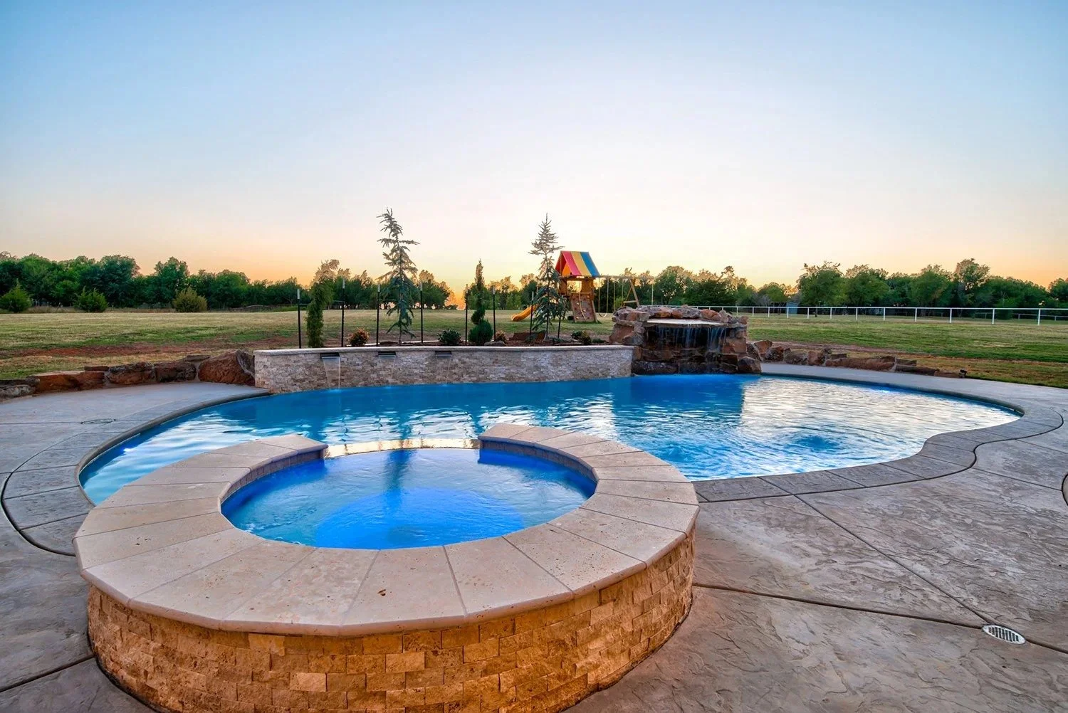 A backyard with a swimming pool and hot tub, with a grassy field, trees, a small playground with a slide, and a sunset sky in the background.
