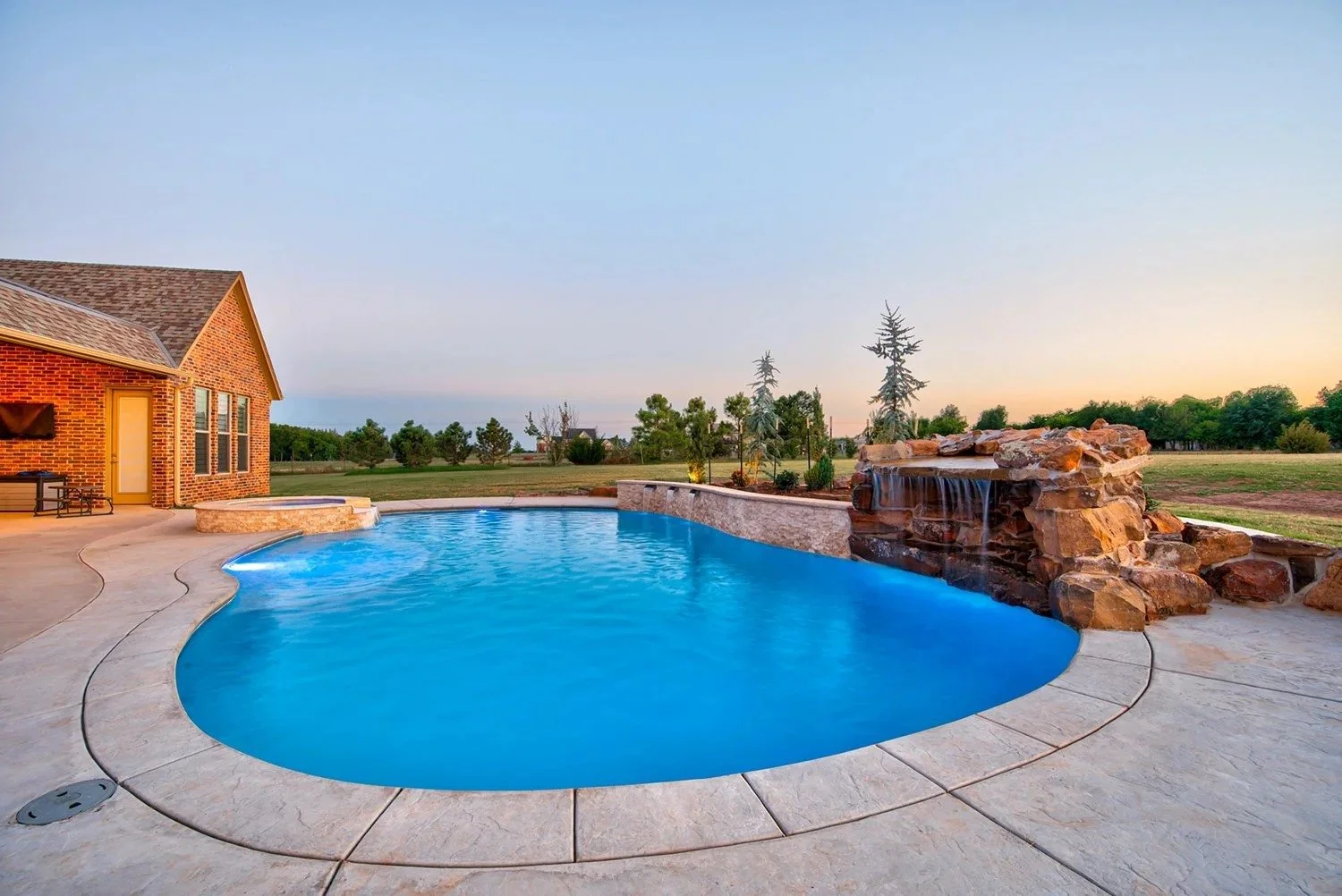 A backyard with a swimming pool, waterfalls, a brick house, and green trees at sunset.