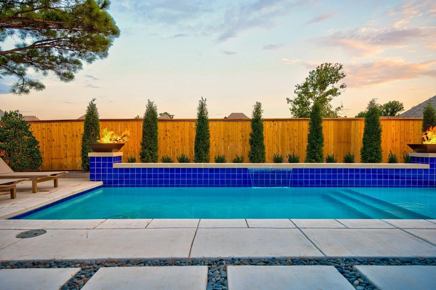 A backyard swimming pool with blue tiles, surrounded by white stone paving, two lounge chairs on the left, a tall wooden fence, several green shrubs and trees, two fire bowls with flames on either side, and a sky with clouds at sunset.