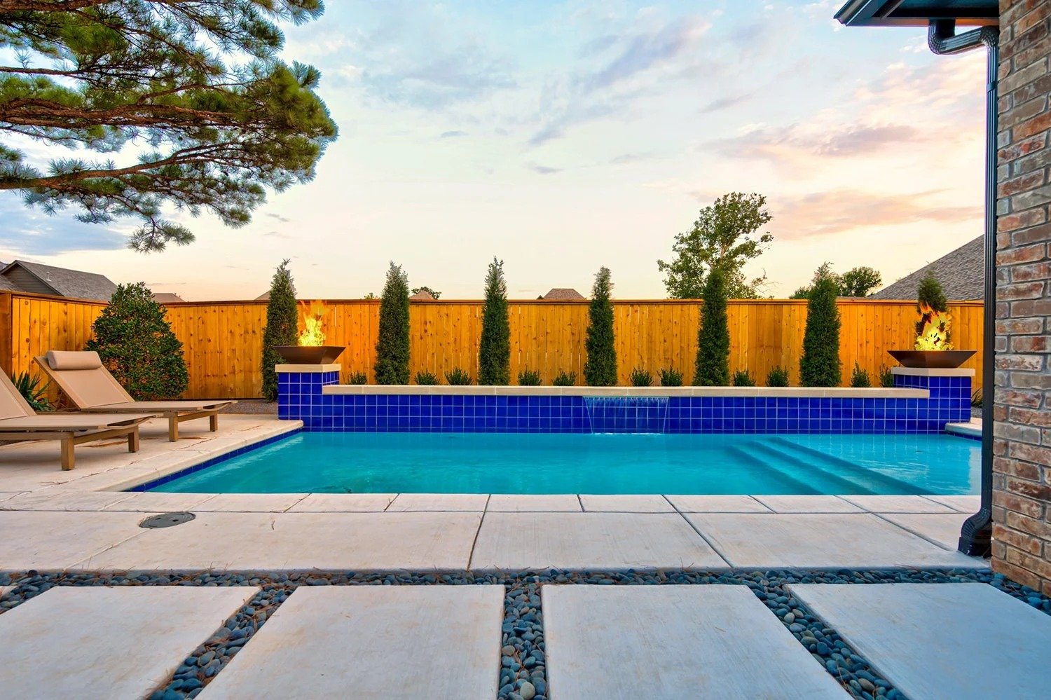 A backyard swimming pool with a blue tile border, two lounge chairs on the left, and a wooden privacy fence with tall green trees and shrubs in the background at sunset.