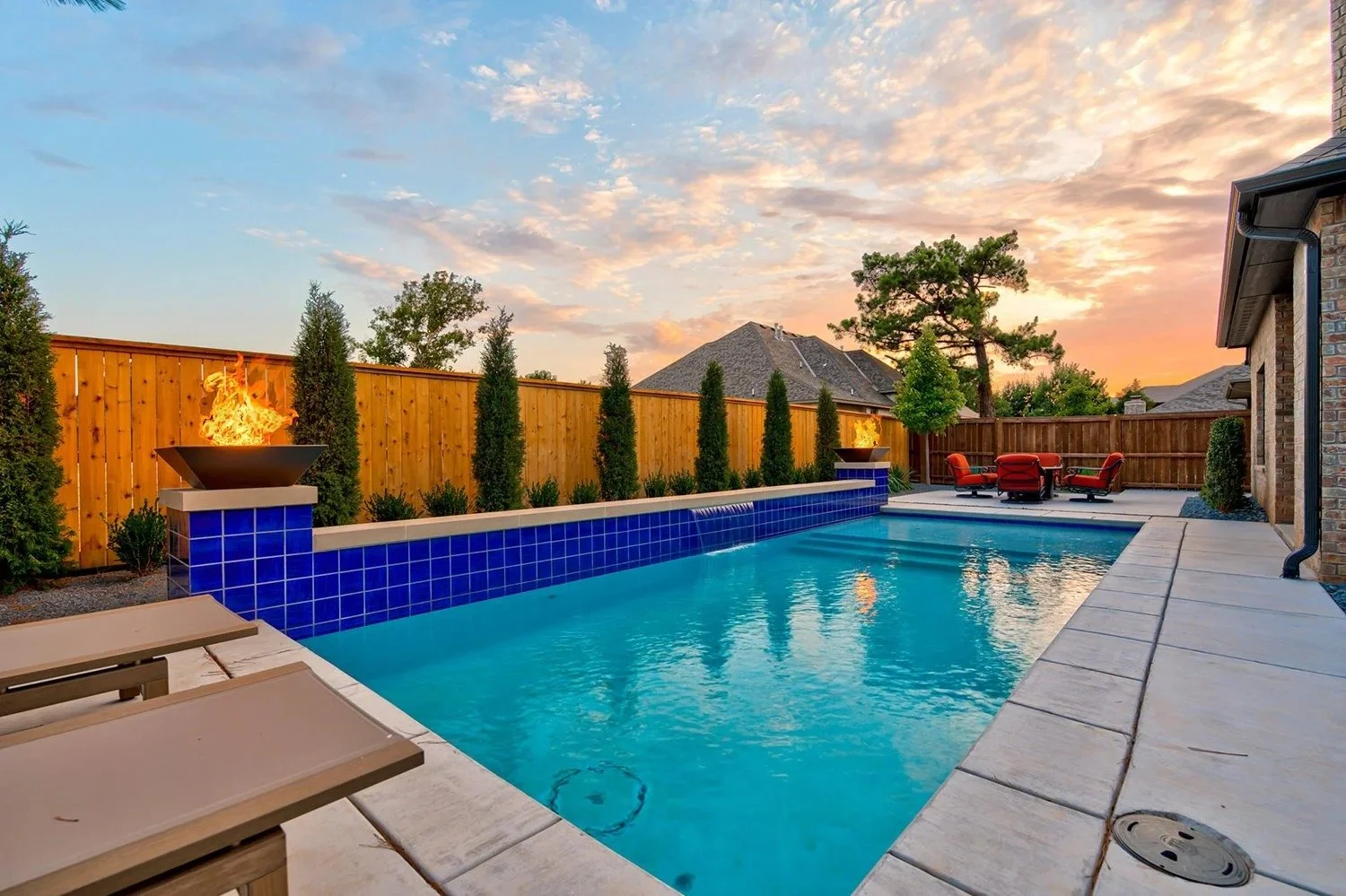 A backyard swimming pool with a blue tile waterfall, surrounded by a concrete deck, with fire features on either side, outdoor seating with three red chairs and a table, a wooden privacy fence, green trees, and a sunset sky.