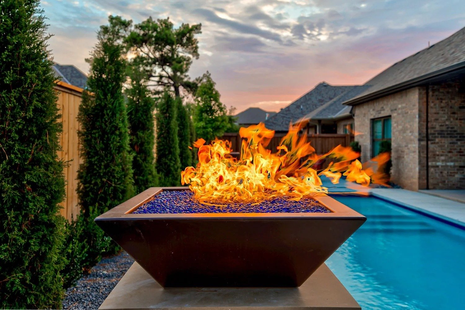A backyard scene during sunset featuring a rectangular fire pit with bright flames, a swimming pool, tall green hedges, and houses with gabled roofs.