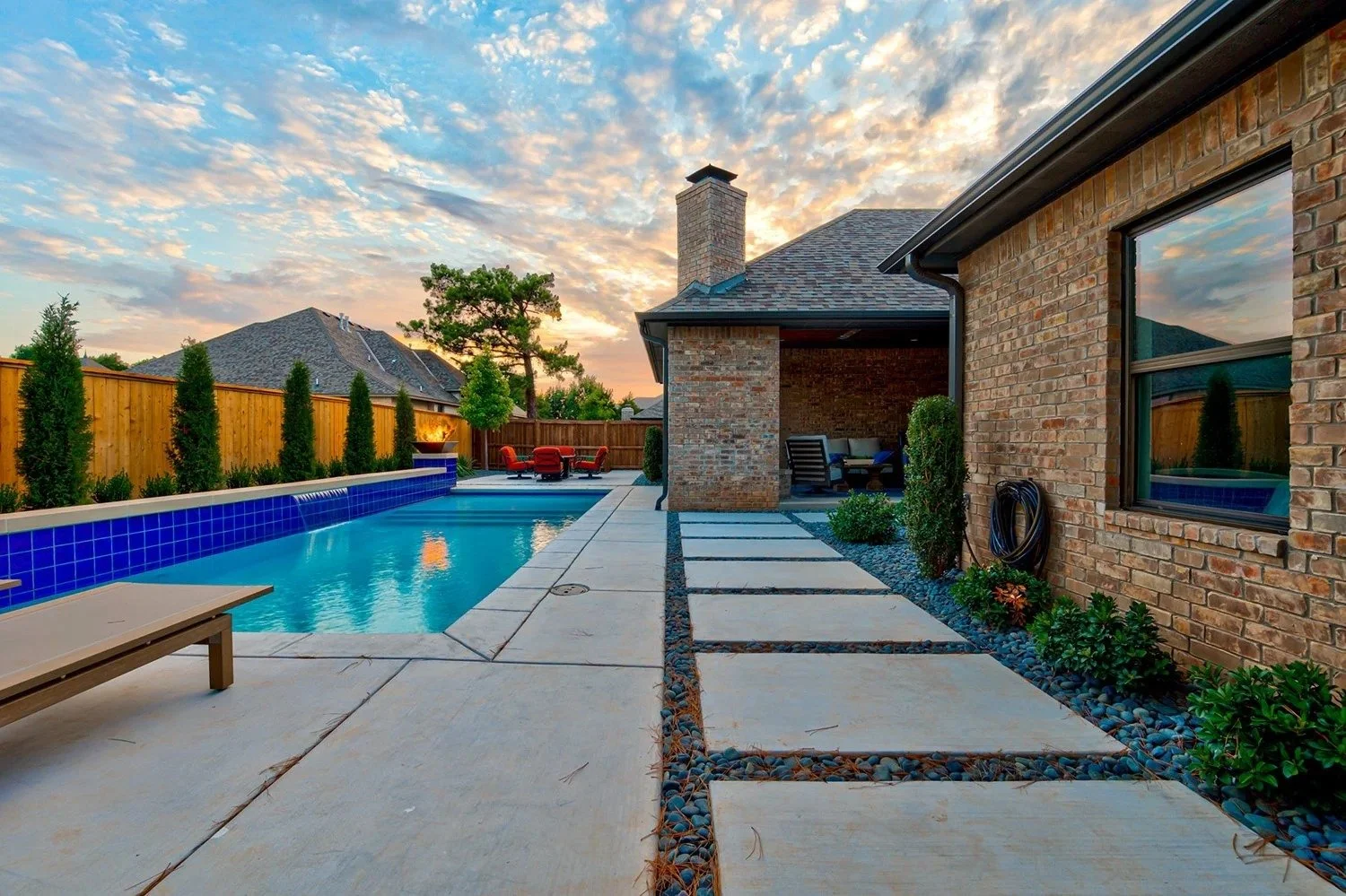 Backyard with swimming pool, patio area, outdoor seating, and a brick house under a sunset sky.