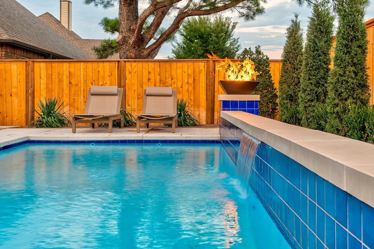Swimming pool with two lounge chairs, a wooden fence, a fire feature next to tall shrubs and trees, and a clear sky in the background.
