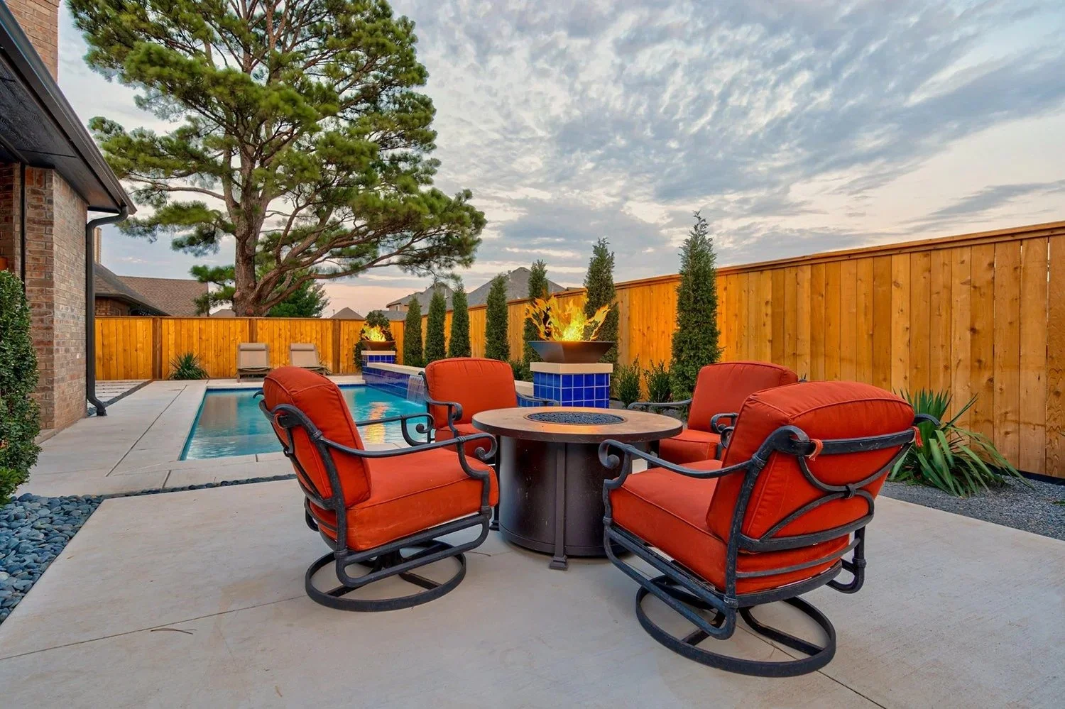 Backyard patio with a pool, wooden fence, and outdoor seating with four red cushioned chairs and a round fire pit table, under a partly cloudy sky.