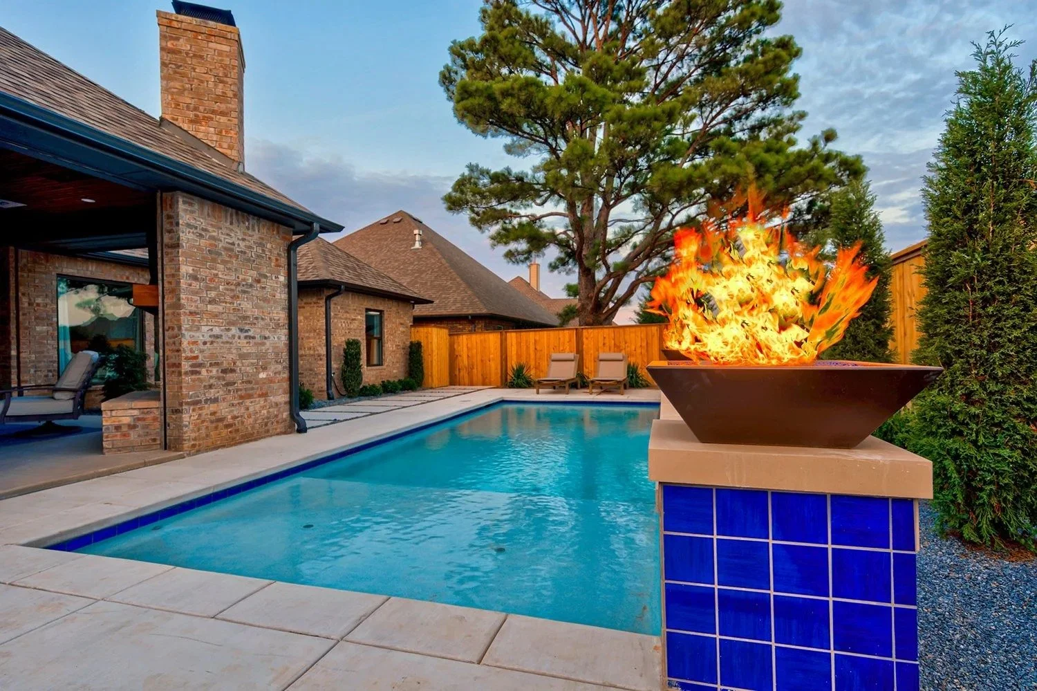 Residential backyard with a swimming pool, a fire feature with flames, a patio area with chairs, and a wooden fence surrounded by trees.