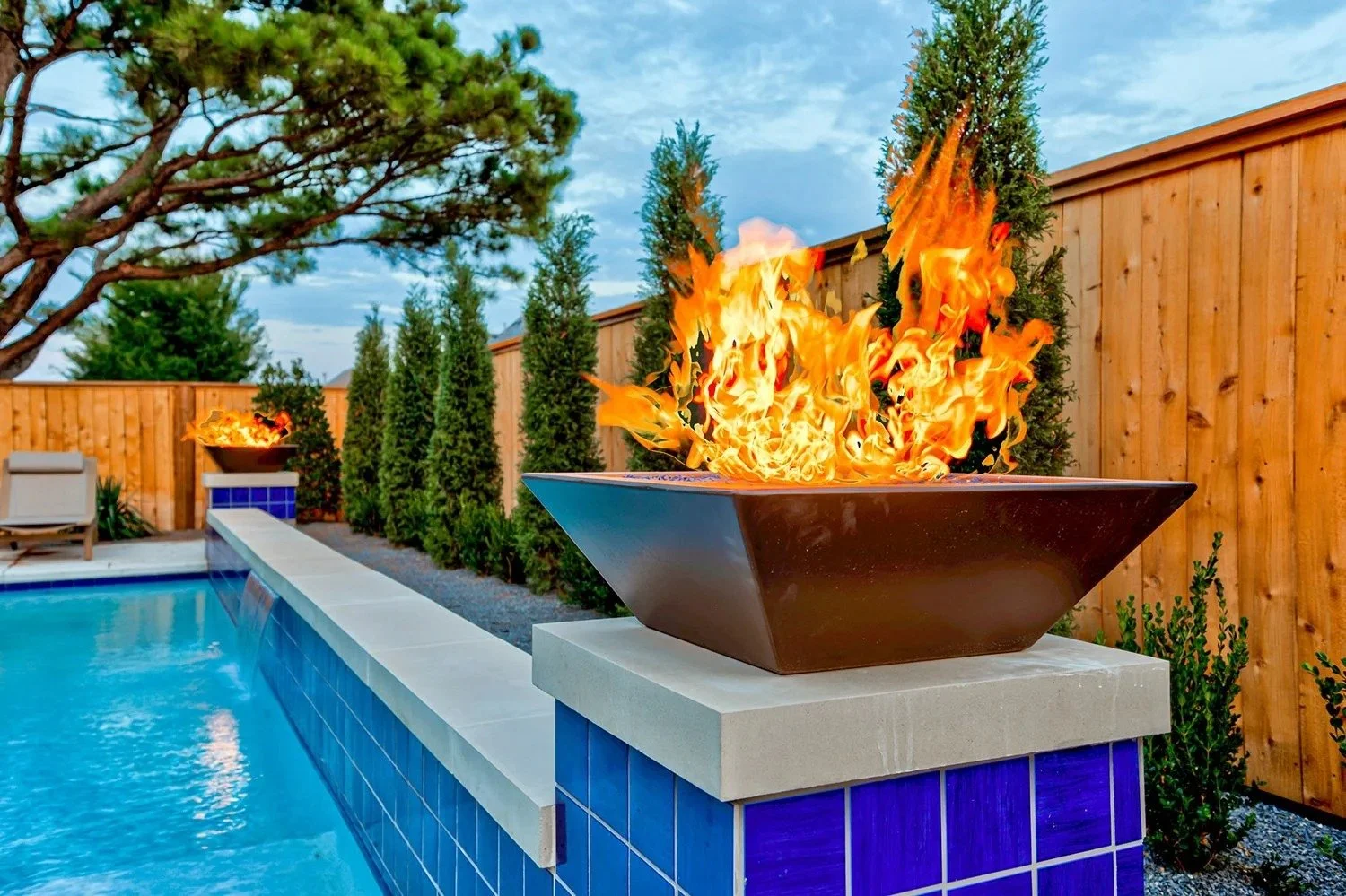 A backyard swimming pool with a fire feature in a square bowl on a square pedestal, with flames rising from it. A wooden fence surrounds the backyard, with tall evergreen trees and a large tree with branches extending over the yard.