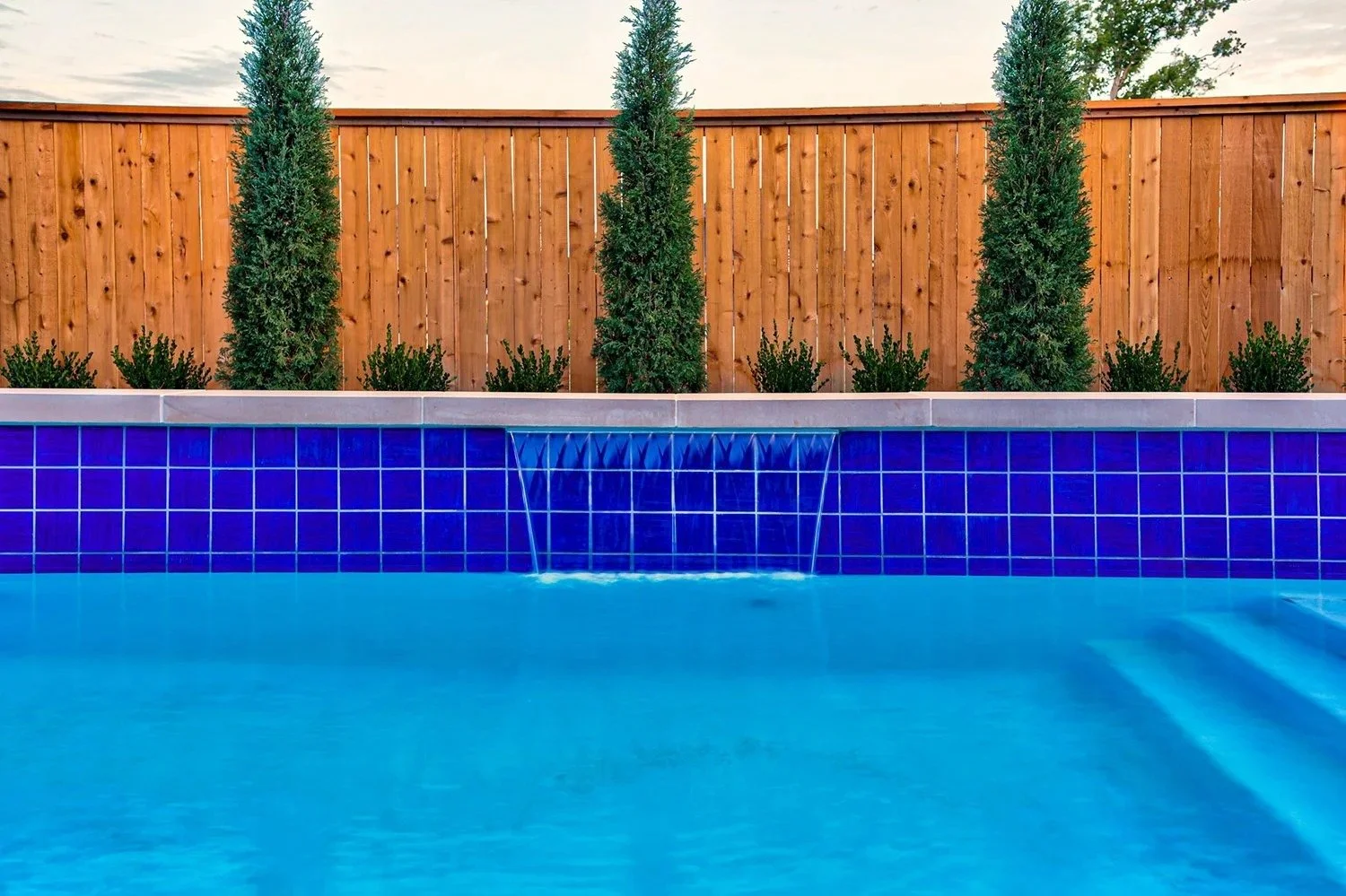 Backyard swimming pool with blue tiles, water feature, and a wooden fence with small trees and shrubs behind it.