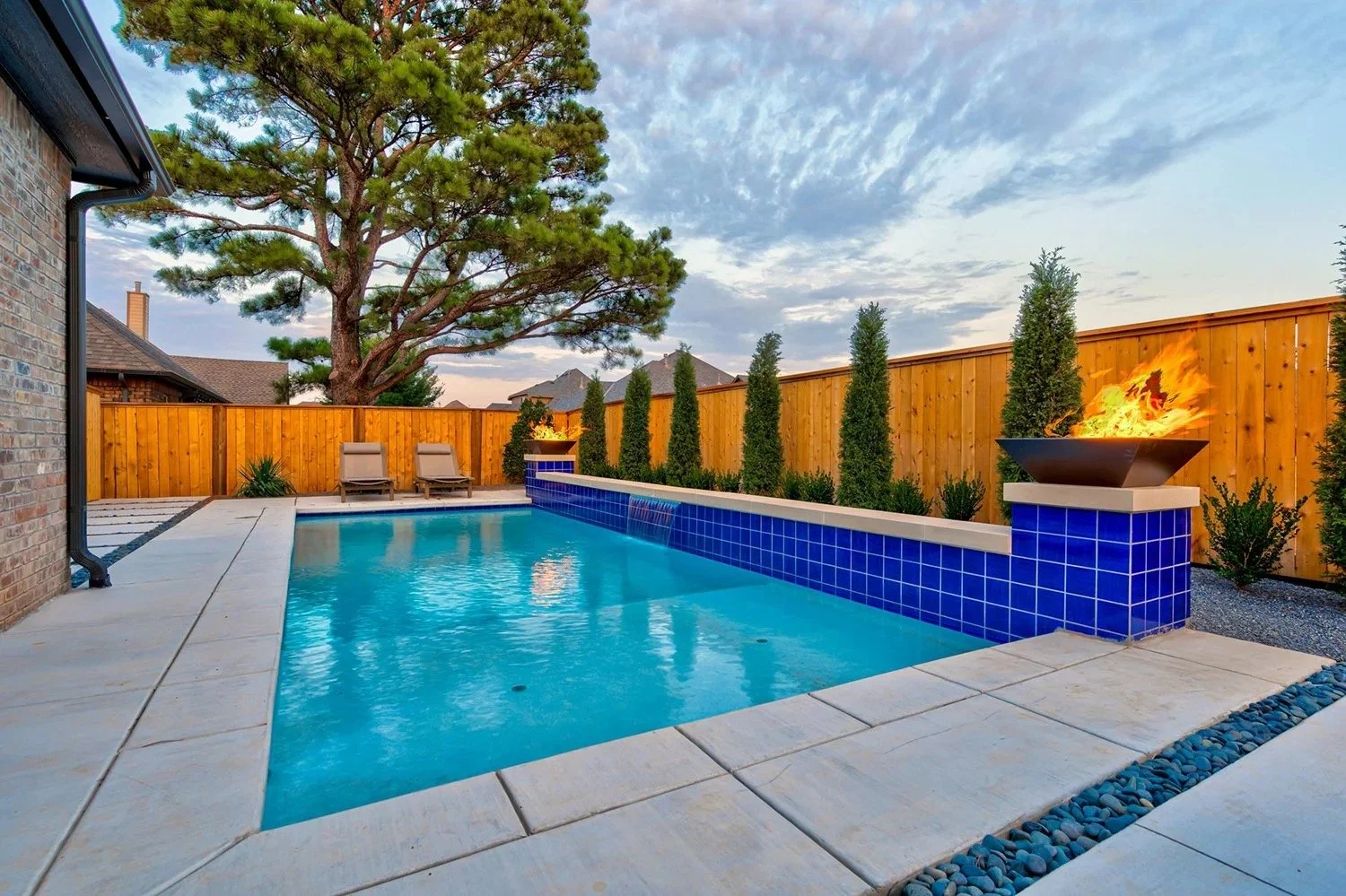 A backyard with a rectangular swimming pool surrounded by a concrete deck, two lounge chairs, a wooden fence with tall shrubs, a large tree, and a fire feature with flames in a modern bowl.