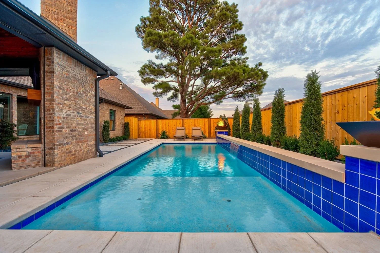 A backyard with a swimming pool, two lounge chairs, a large tree, and a wooden fence at sunset.