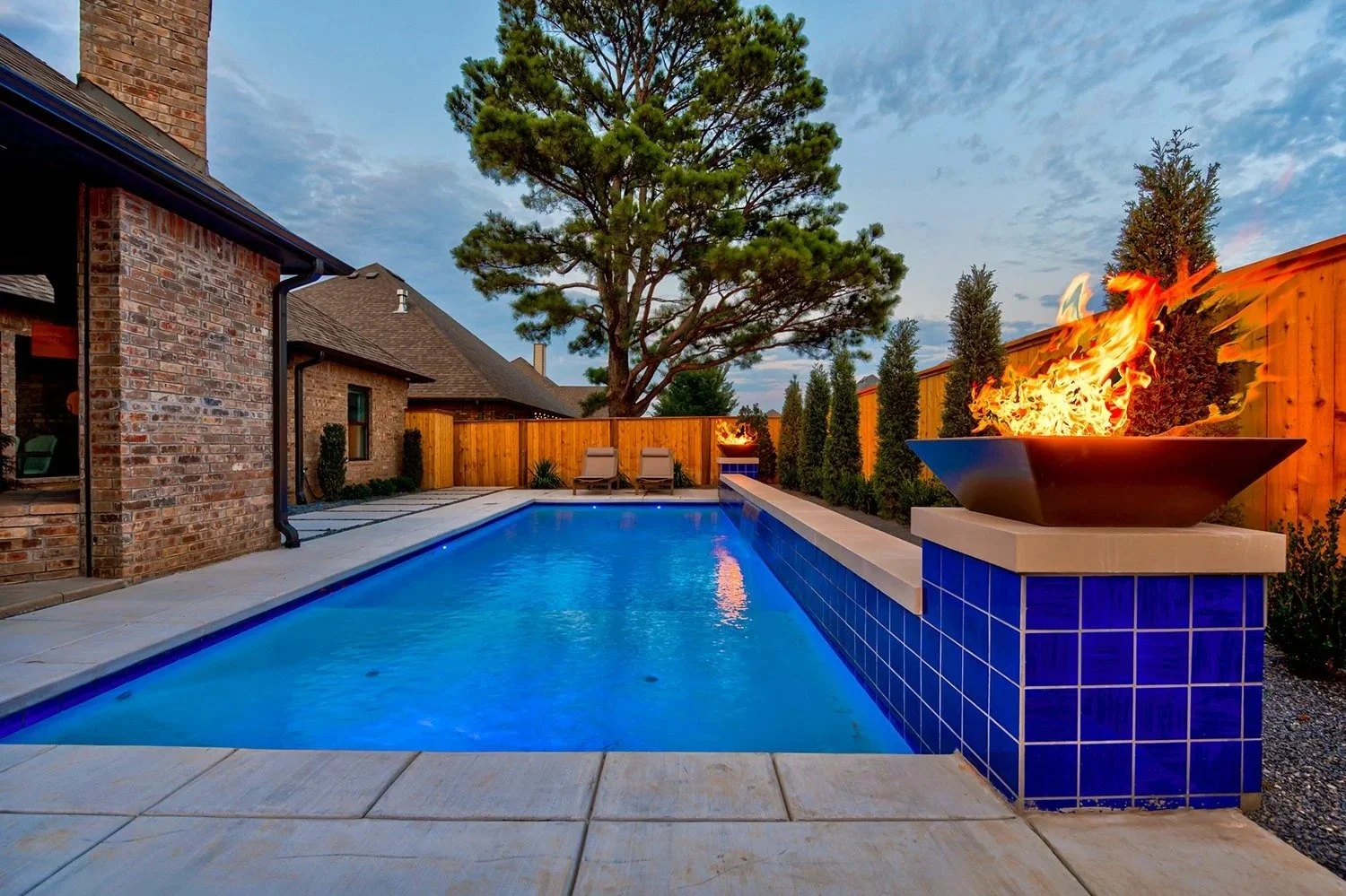 A backyard swimming pool with blue water, surrounded by a concrete deck and a wooden privacy fence. There are two lounge chairs at the far end of the pool, and a fire pit with a burning flame on a tiled pedestal. Tall trees and a brick house are in t