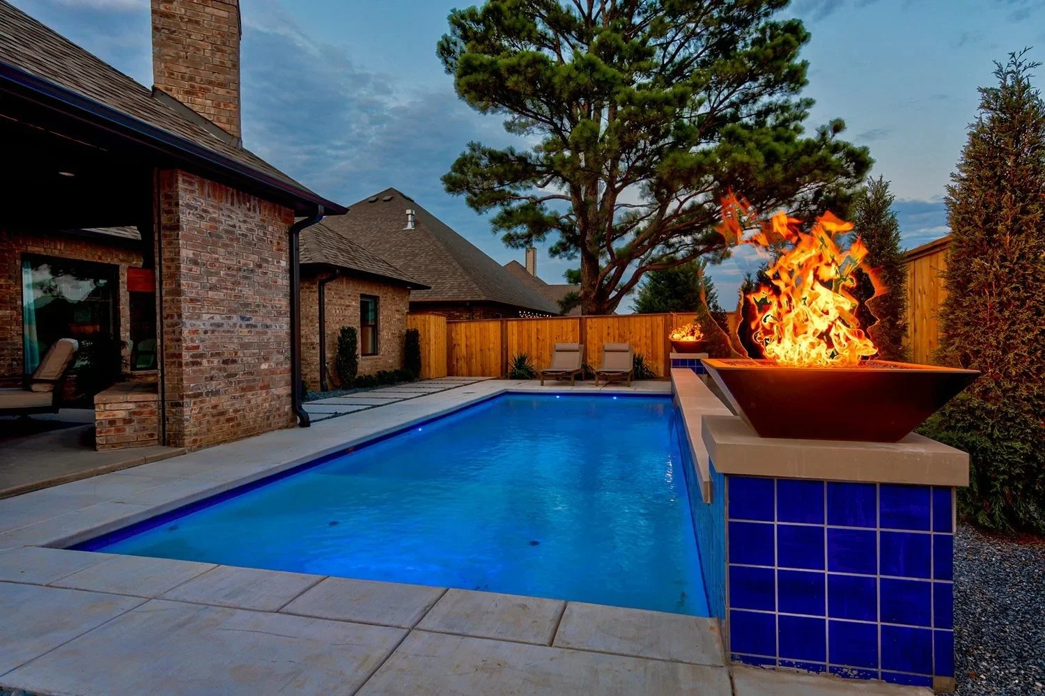 Backyard with a swimming pool, a fire feature with flames, lounge chairs, a wooden fence, trees, and a brick house in the evening.