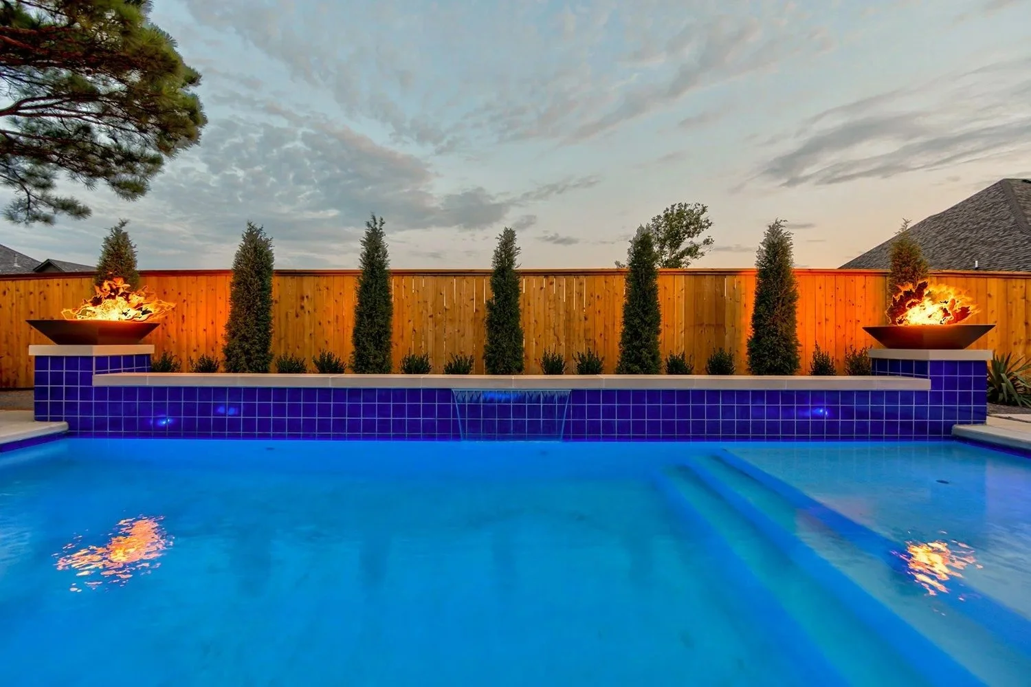 A backyard swimming pool with blue tiles, two fire features on either side, a wooden privacy fence, tall trees, and a sunset sky.