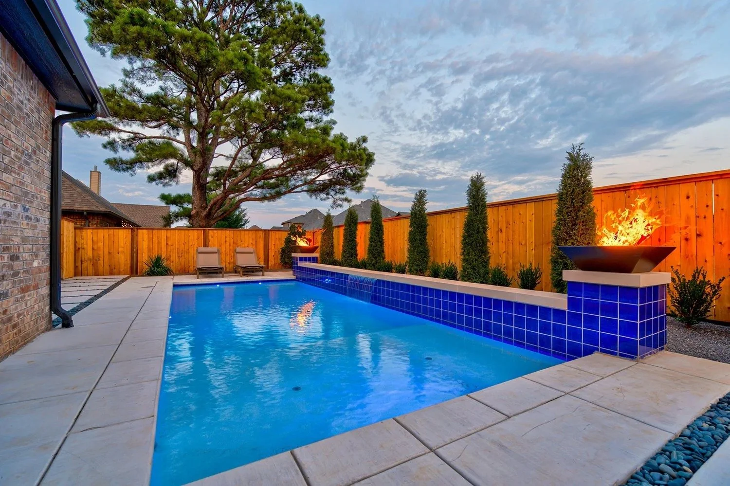 A backyard with a rectangular swimming pool with blue tile border, two lounge chairs, a large tree, decorative plants, and a fire feature on a blue tiled pedestal during dusk.