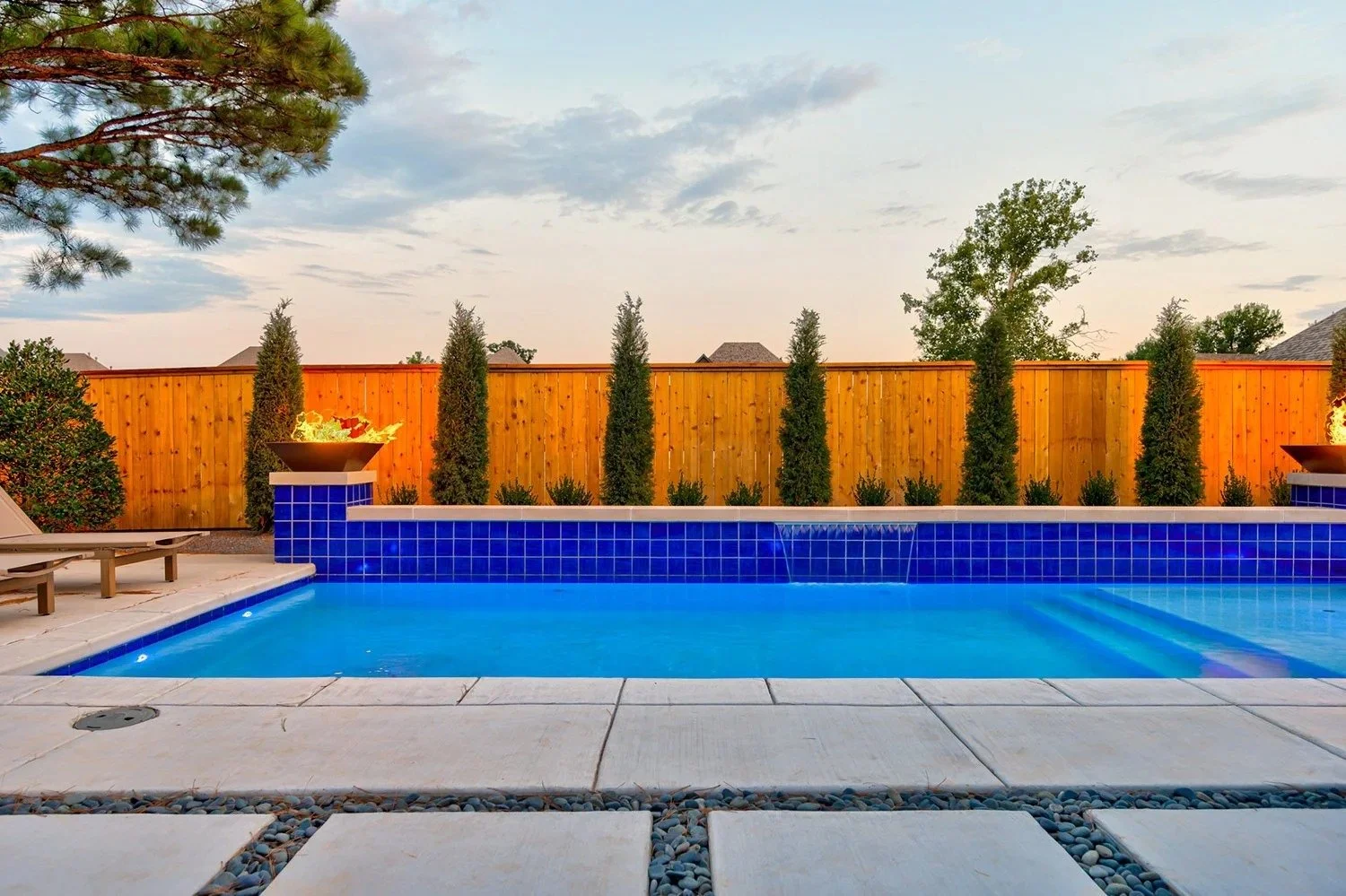 A backyard swimming pool with blue tiles, surrounded by a white stone patio and black pebbles. A wooden privacy fence with tall, narrow trees and decorative fire bowls on blue tiled pedestals are visible in the background. There are lounge chairs on 