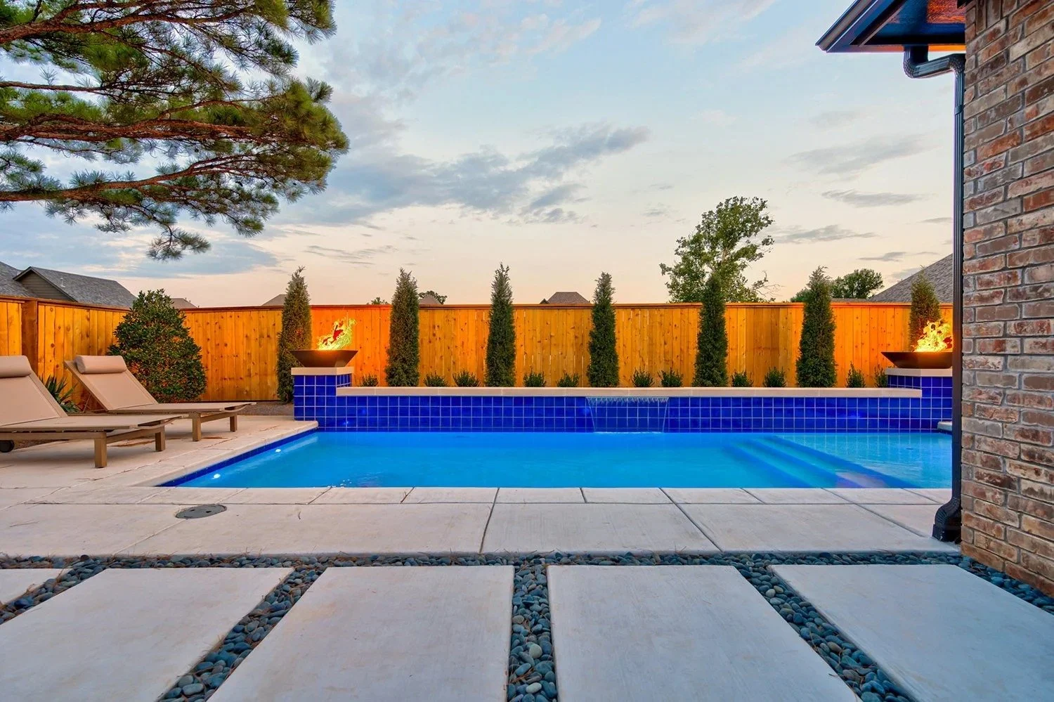 A backyard with a swimming pool, poolside lounge chairs, and a wooden fence with trees and shrubs behind it. The pool has a blue tiled edge with water features and two fire bowls with flames.