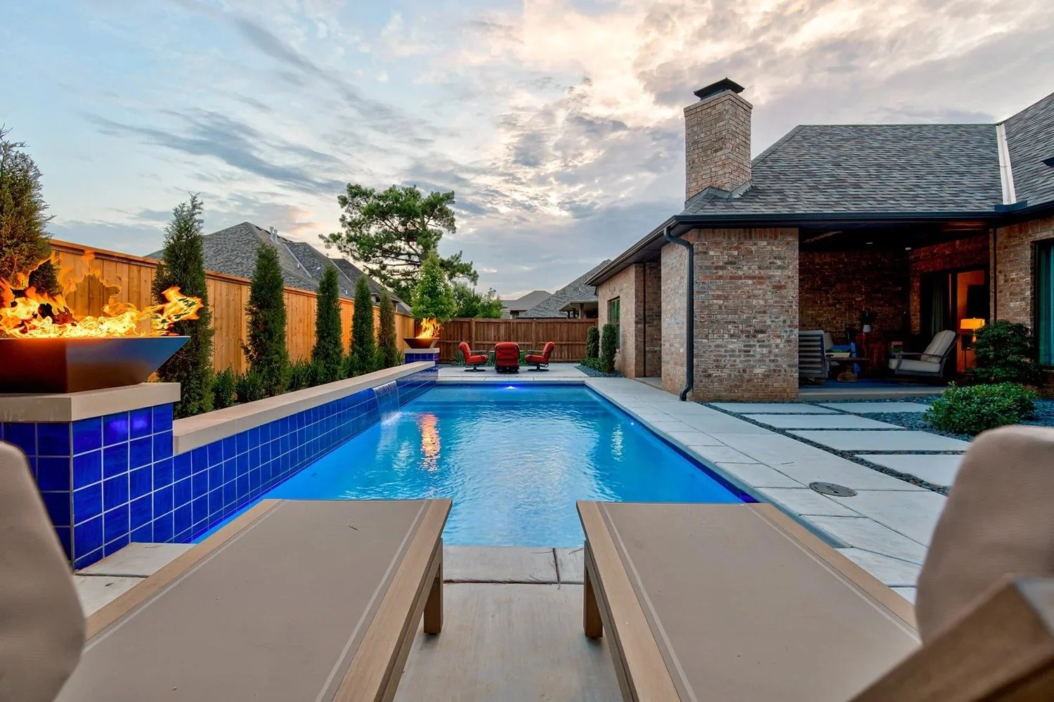 A backyard with a swimming pool, blue tiles, and a fire feature at each end. There are lounge chairs in the foreground and patio furniture with red cushions at the far end. The backyard is enclosed by a wooden fence with greenery and trees, and a hou