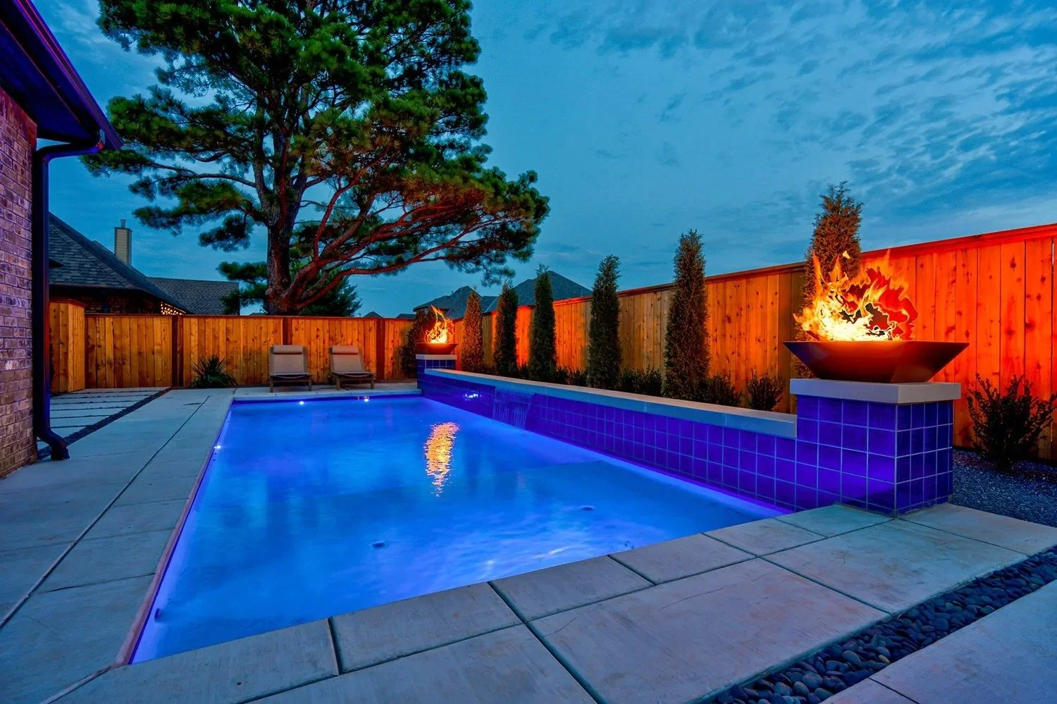A backyard swimming pool at dusk with blue lighting, surrounded by a wooden fence. Fire bowls with flames are on pedestals along the fence, and two lounge chairs are positioned near a large tree.