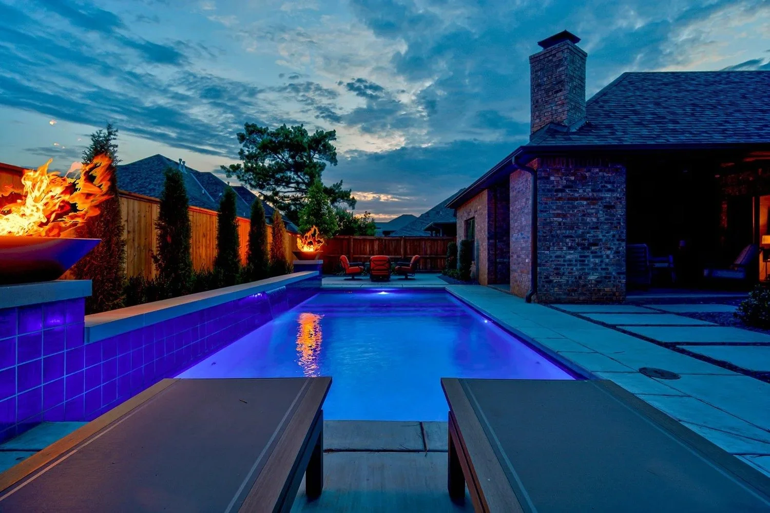 Backyard swimming pool illuminated with blue lights at dusk, surrounded by a wooden fence, with fire features on either side, patio furniture in the background, and a brick house nearby.