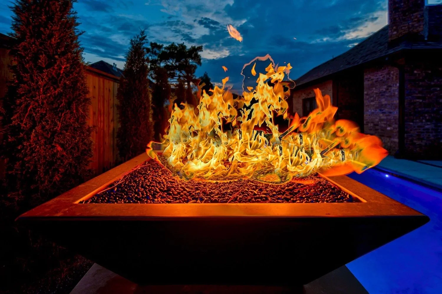 Burning fire in an outdoor fire pit on a backyard deck, with a fence, trees, and a house in the background during dusk.