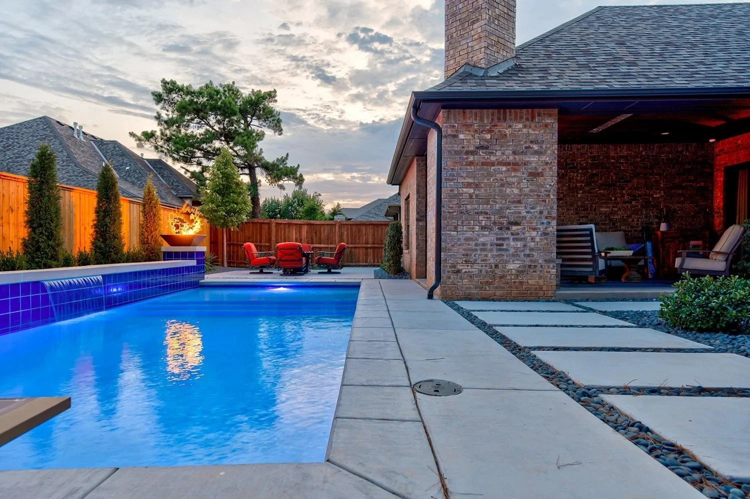 Residential backyard with a swimming pool, outdoor furniture, and a fire feature. The yard is enclosed with a wooden fence, and the house has brick exterior walls with a covered patio area.
