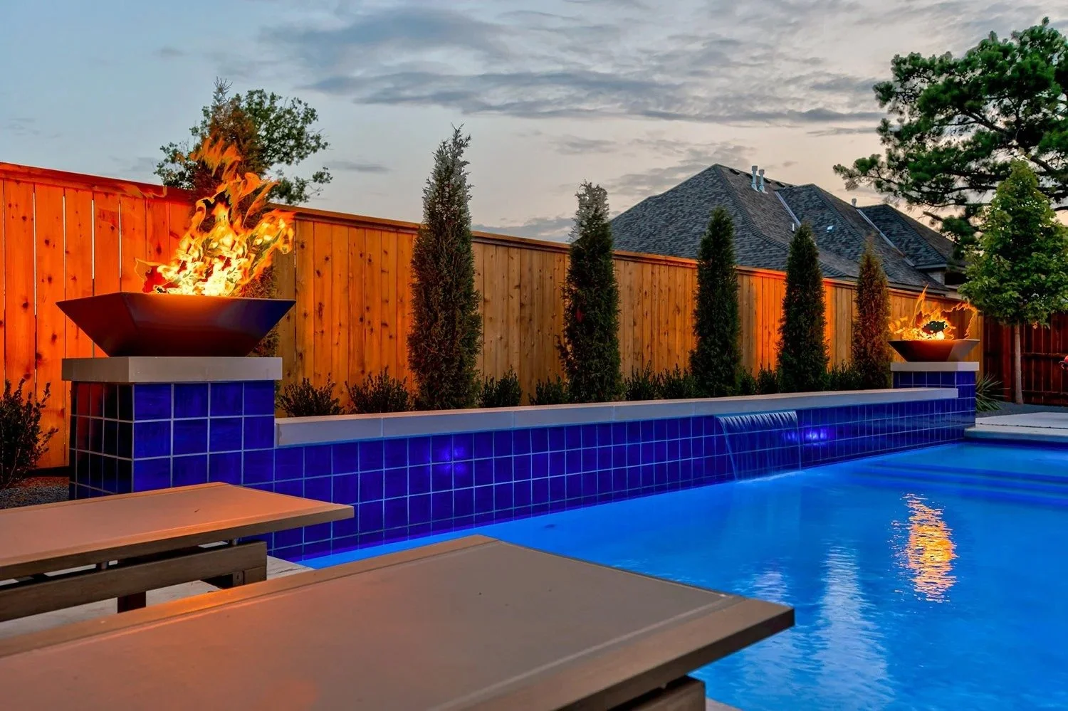 A backyard swimming pool with a blue tile border, lit by two flames in decorative fire bowls on either side, surrounded by wooden fencing and tall trees at dusk.