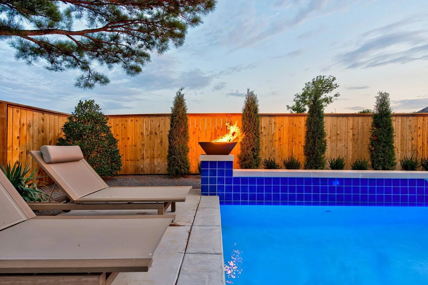 A backyard swimming pool with blue tiles, two beige lounge chairs, a fire pit with flames, a wooden fence, and tall evergreen trees under a partly cloudy sky at sunset.
