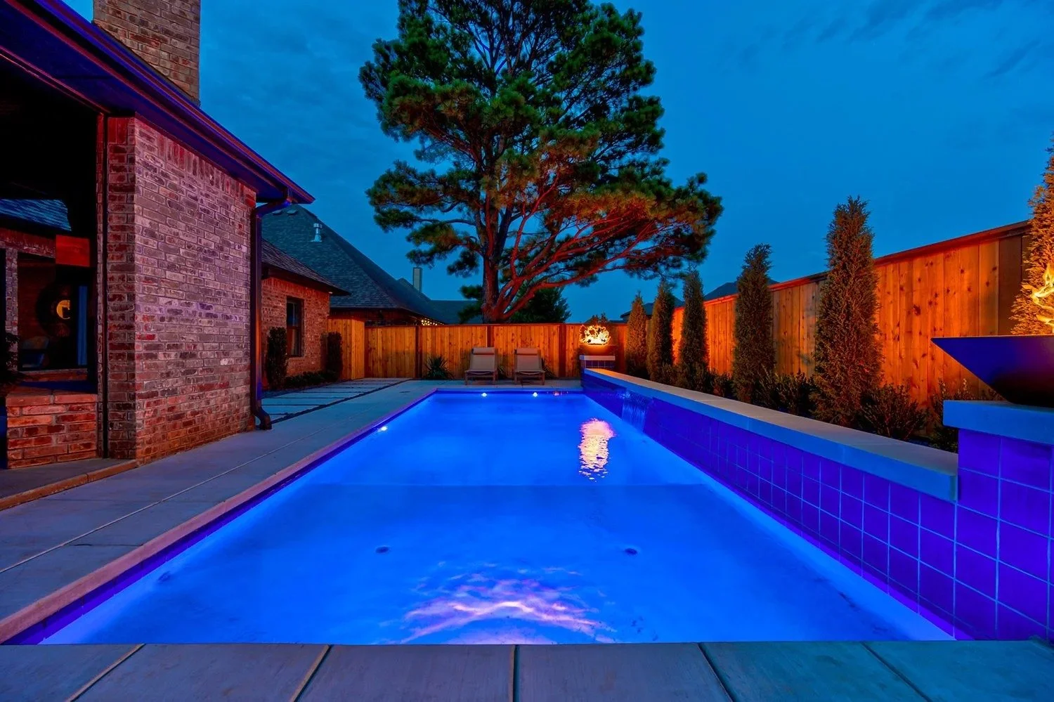 Backyard swimming pool illuminated with blue lights during dusk, surrounded by a wooden fence, with two lounge chairs at the far end, a large tree in the background, and a brick house on the left side