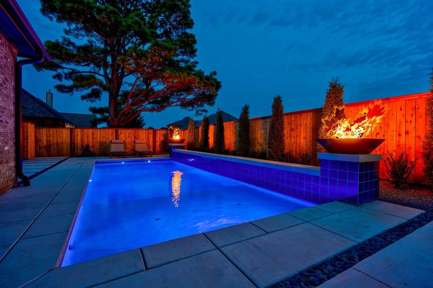 A backyard swimming pool at dusk with blue lighting, a wooden privacy fence, a large tree, two lounge chairs, and fire features on either side.