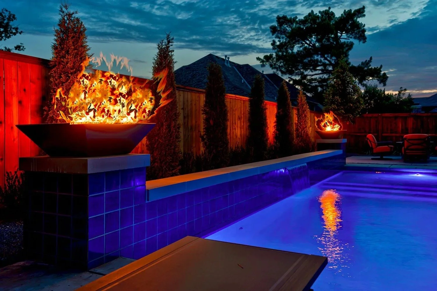 A backyard at dusk featuring a swimming pool with blue lighting, two fire bowls with flames on either side, a wooden fence with tall trees, and outdoor seating area.